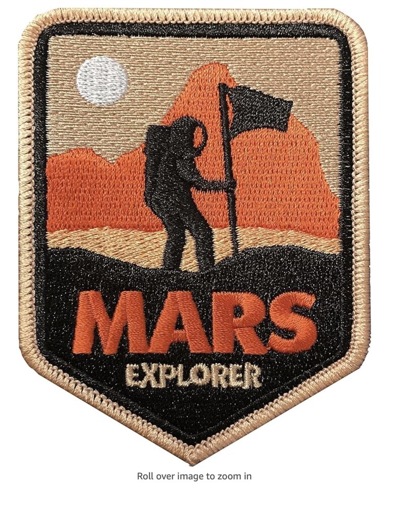 Puede incluir: Un parche bordado con una silueta negra de un astronauta plantando una bandera en una cordillera roja y naranja. El texto "MARS EXPLORER" est&aacute; debajo de la imagen en letras naranja y blancas sobre un fondo negro.