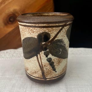 May include: A cylindrical ceramic cup featuring a speckled cream-coloured base with dark brown horizontal bands at the top and bottom. The exterior is decorated with a dark brown painted dragonfly motif, showcasing detailed wings and a segmented body.