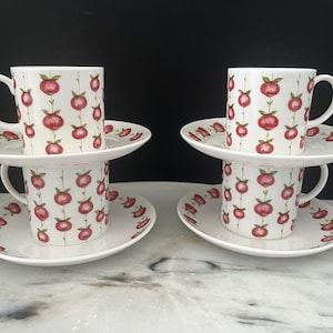 May include: Two sets of white ceramic cups and saucers featuring a repeating pattern of red pomegranate-like fruits with green stems and leaves. Each cup has a curved handle, and the saucers are decorated with the same fruit motif along the rim.
