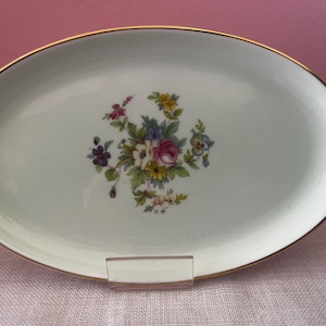 May include: An oval white porcelain serving platter featuring a central floral bouquet with pink, yellow, blue, and purple flowers surrounded by green leaves. The platter has a thin gold rim around the edge and a smooth, glossy finish.