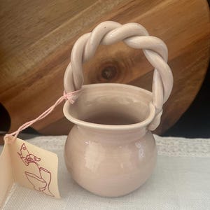 May include: A small, light pink ceramic basket with a smooth, glossy finish and a twisted, rope-style handle. A light pink string is attached to the handle, holding a rectangular paper tag featuring a line drawing of a butterfly and a vessel.