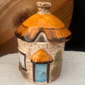 May include: A ceramic jar shaped like a cottage with a removable lid featuring a textured orange thatched roof and a chimney. The base depicts a cream-coloured brick wall, a bright blue door, a window with a grid pattern, and green foliage.