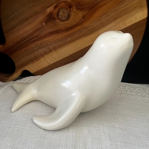 May include: A smooth, off-white ceramic figurine of a seal in a resting position. The seal is depicted with its head tilted upward, featuring a streamlined body, a rounded snout, and a bifurcated tail fin, all rendered in a glossy, solid finish.