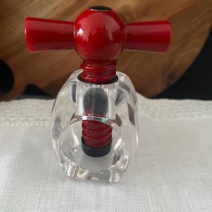 May include: A corkscrew featuring a clear acrylic body with a red handle and a red internal screw mechanism. The design includes a black circular top accent and a black base component, showcasing a transparent structure with bold red and black accents.