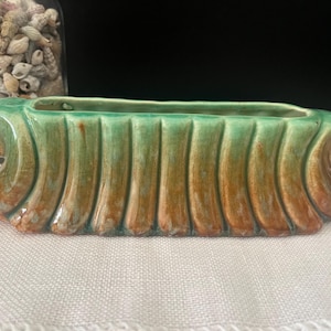 May include: An elongated ceramic planter featuring a vertical ribbed texture with a gradient glaze transitioning from a soft sage green at the top to a warm, earthy brown at the base, finished with curled, scroll-like ends on both sides.
