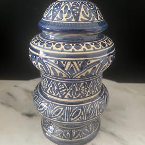 May include: A blue and cream ceramic jar with a lid. The jar has detailed patterns in cream. The jar is a decorative item. The height of the jar is approximately 20 cm.