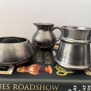 May include: Three pewter-coloured metal containers: a small bowl, a creamer jug, and a handled mug. The objects are arranged on a book with the title "ANTIQUES ROADSHOW" visible. The items have a vintage aesthetic.