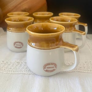 May include: A set of six ceramic mugs featuring a white base and a glossy tan rim. Each mug has a handle and a circular stamp on the front that reads Bendigo Pottery Heritage Australia. The mugs are arranged in a group on a white lace surface.