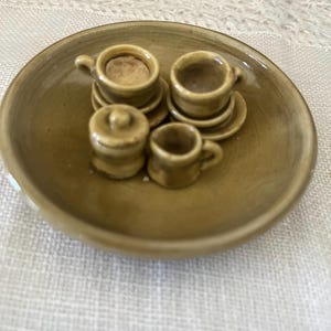 May include: A miniature ceramic tea set in a glossy olive-brown glaze, consisting of a round serving tray, two small cups on saucers, a lidded sugar bowl, and a small creamer pitcher, all arranged together as a single cohesive tabletop display.