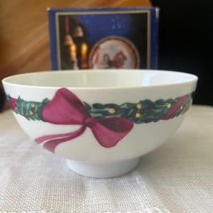 May include: A white porcelain bowl featuring a decorative band around the exterior. The design consists of a green holly garland with small gold dots and a prominent dark pink ribbon tied in a bow. The bowl has a smooth, rounded shape.