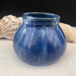 May include: A small, round, blue ceramic vase with a glossy finish. The vase has a wide opening and a textured rim. The blue colour has white streaks throughout. The vase is on a white surface.