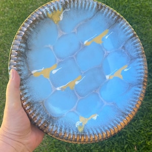 May include: A round ceramic plate with a light blue, textured surface and a gold rim. The plate has a unique pattern of interconnected shapes. The plate is held by a hand against a green background.