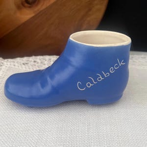 May include: A small, blue ceramic shoe-shaped ornament with a smooth, matte finish. The word Caldbeck is written in white cursive script on the side of the shoe. The interior of the shoe is white, and the overall design is simple and functional.