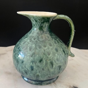 May include: A ceramic pitcher featuring a rounded body and a curved handle. The exterior displays a mottled, variegated glaze in shades of deep forest green and lighter sage green, creating a textured, cloud-like pattern across the surface.