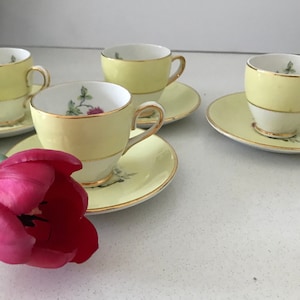 May include: Four vintage teacups and saucers. The cups and saucers are pale yellow with gold edging. Each cup has a white base and a floral design inside. A vibrant red tulip is in the foreground.