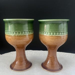 May include: Two ceramic goblets featuring a two-tone design with a glossy green upper section and a speckled brown lower section. A decorative band of raised dots separates the two colors on the bowl, which sits on a sturdy pedestal base.