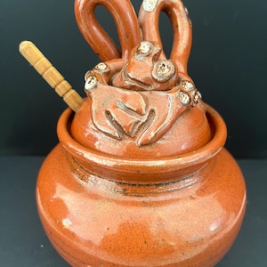 May include: A terracotta-coloured ceramic jar with a lid and a small wooden spoon. The lid features a decorative frog design with raised details. The jar has a round body and a textured surface, ideal for storing spices or other small items.