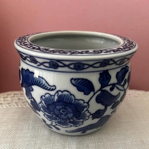 May include: A small, rounded ceramic bowl featuring a white base with intricate blue floral patterns and a decorative border around the rim. The design includes detailed blossoms and leafy vines painted in a deep blue hue against the white surface.