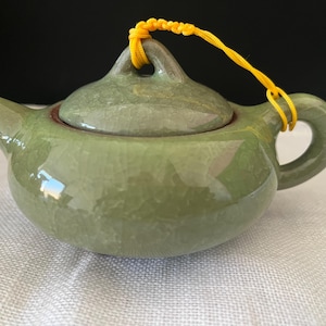 May include: A small, sage green ceramic teapot with a glossy, crackled glaze finish. The teapot features a rounded body, a short spout, a matching lid with a central handle, and a curved side handle attached to a bright yellow cord loop.