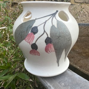 May include: A white ceramic vase with a wide, flared rim and a rounded body featuring two circular cutouts near the top. The surface is decorated with a painted botanical motif of grey leaves, dark grey buds, and soft pink flower stamens.