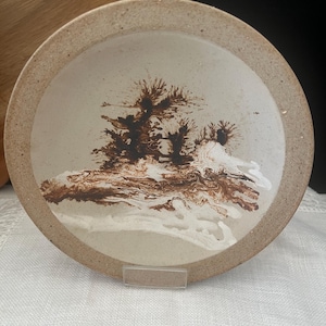May include: A round ceramic plate with a speckled beige rim and a smooth off-white centre. The plate features a dark brown, tree-like dendritic pattern in the middle, accented by swirling white glaze details that resemble a landscape scene.