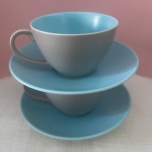 May include: Two stacked tea cups and saucers featuring a two-tone design. The exterior of each cup and the handle are a solid matte grey, while the interior of the cup and the top surface of the saucer are a vibrant, smooth light blue finish.
