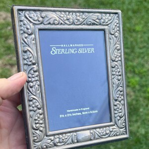 May include: A rectangular sterling silver picture frame with intricate floral patterns. The frame is marked "HALLMARKED STERLING SILVER" and "Handmade in England". The frame measures 3.5 x 2.5 inches, and 9cm x 6.5cm.