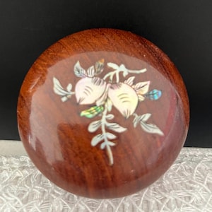 May include: A round, polished wooden box lid featuring a central inlay of iridescent mother-of-pearl. The inlay depicts a botanical design with two rounded fruit-like shapes and leafy stems, showcasing shimmering white, pink, and green tones.