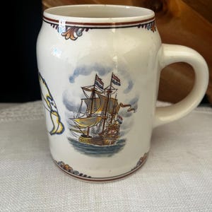 May include: A cream-coloured ceramic mug featuring a detailed illustration of a historic sailing ship on the water. The mug has a rounded body, a sturdy handle, and decorative brown and blue floral-style trim along the top and bottom rims.