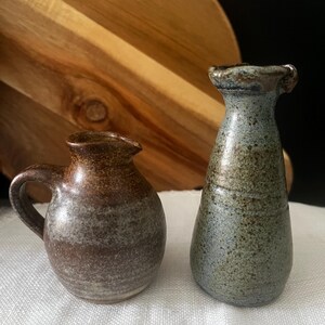 May include: A small, round, brown ceramic jug with a handle and a narrow spout, positioned next to a taller, slender, blue-green ceramic vase with a flared rim. Both vessels feature a speckled, textured glaze finish on their surfaces.