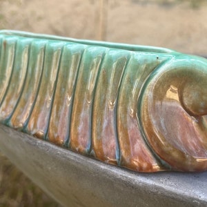 May include: A ceramic planter with a green, brown and gold glaze. The planter has a wavy, organic shape and a ribbed texture.