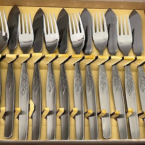 May include: A set of ten silver-toned forks and knives with decorative etched designs. The utensils are arranged in a box with a beige interior.