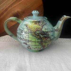 May include: A round ceramic teapot featuring a light green and blue world map design. The surface displays printed geographical labels including Mer Pacifique and Mer du Nord, with a matching lid, a curved handle, and a tapered spout for pouring.