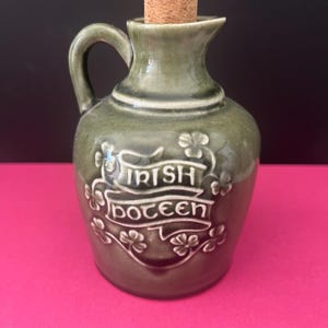 May include: A small, green ceramic jug with a handle and a cork stopper. The jug is embossed with the words "Irish Poitín" and shamrock designs. The jug is on a pink surface with a dark background.