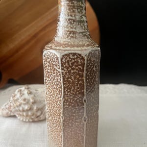 May include: A tall, hexagonal ceramic bottle with a textured, speckled brown and cream glaze. The bottle features a narrow neck and a distinct vertical panel design, showcasing a mottled, earthy finish across its entire surface.