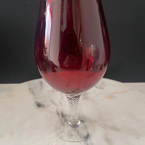 May include: A large, burgundy-coloured wine glass with a clear, twisted stem and a wide bowl. The glass is set against a marble surface and a dark background, creating a contrast that highlights its shape and colour.