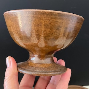 May include: A brown ceramic sundae dish with a pedestal base. The dish has a rounded bowl shape and a textured surface. The color is a warm, earthy brown with lighter highlights. The dish is held in a hand.