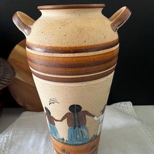 May include: A speckled tan ceramic vase with two handles, featuring horizontal brown bands and a hand-painted scene of three figures holding hands in blue skirts and brown tops, with small green grass details near the base and a black bird above.