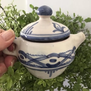 May include: A small ceramic sugar bowl with a matching lid, featuring a cream-coloured base and hand-painted blue decorative patterns. The bowl has two side handles and a small spout, with intricate blue line work encircling the main body.