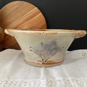 May include: A ceramic bowl with two small handles, featuring a speckled off-white glaze with a central blue and purple abstract brushstroke design. The bowl has a wide, flared rim and a sturdy base with visible brown clay tones.