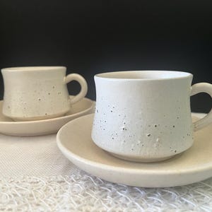 May include: Two off-white ceramic cups with matching saucers, featuring a speckled texture throughout the surface. Each cup has a rounded body, a small base, and a curved handle. The ceramic has a matte finish with visible dark flecks.