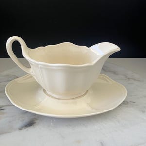 May include: Cream-coloured gravy boat and matching saucer. The boat has a curved handle and a spout for easy pouring. The set is ceramic and has a scalloped edge design, ideal for serving sauces and gravies.