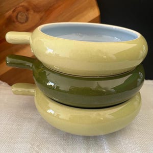 May include: A stack of three ceramic bowls with handles, featuring a glossy finish. The top and bottom bowls are a light sage green, while the middle bowl is a darker forest green. Each bowl has a smooth, rounded rim and a light blue interior.
