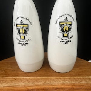 May include: Two white ceramic salt and pepper shakers featuring a printed emblem with the text Compliments WA Branch, The Rats of Tobruk Association, 33rd Anniversary, Adelaide 1974. Each shaker has a smooth, rounded, tapered cylindrical shape.