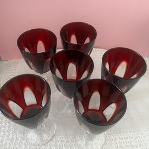 May include: A set of five stemmed glasses featuring deep red glass bowls with clear, vertical, petal-shaped cutouts around the sides. Each glass has a clear glass stem and base, showcasing a contrast between the dark red and transparent glass.