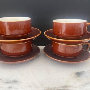May include: Six brown ceramic cups and saucers are stacked in pairs. The cups have a rounded shape with a handle and a white interior. The saucers are round and match the color of the cups. The set is arranged on a white and gray surface.