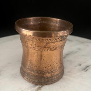 May include: A small, cylindrical copper cup with a flared rim and base. The exterior surface features two horizontal bands of engraved, repeating diagonal hatch marks encircling the body, with a smooth, weathered copper finish throughout.