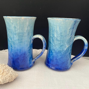 May include: Two ceramic mugs featuring a vertical ombre colour gradient that transitions from a light, pale blue at the top rim to a deep, saturated royal blue at the base. Each mug has a smooth, glossy finish and a curved handle on the side.
