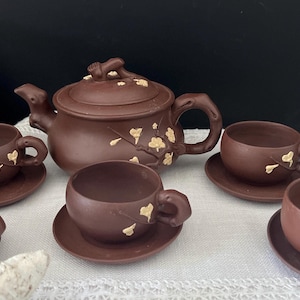 May include: A brown clay tea set consisting of a teapot and four matching cups with saucers. The teapot and cups feature a textured branch design with small, light-coloured floral accents. The teapot lid has a sculpted handle detail.