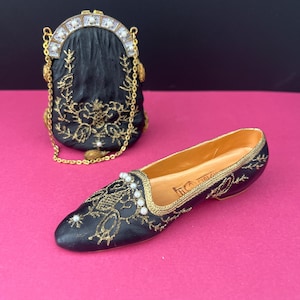 May include: A black miniature handbag with gold embroidery, a gold chain strap, and a clasp featuring white beads, paired with a matching black flat shoe featuring gold embroidery, white bead accents, and a smooth gold-toned interior lining.